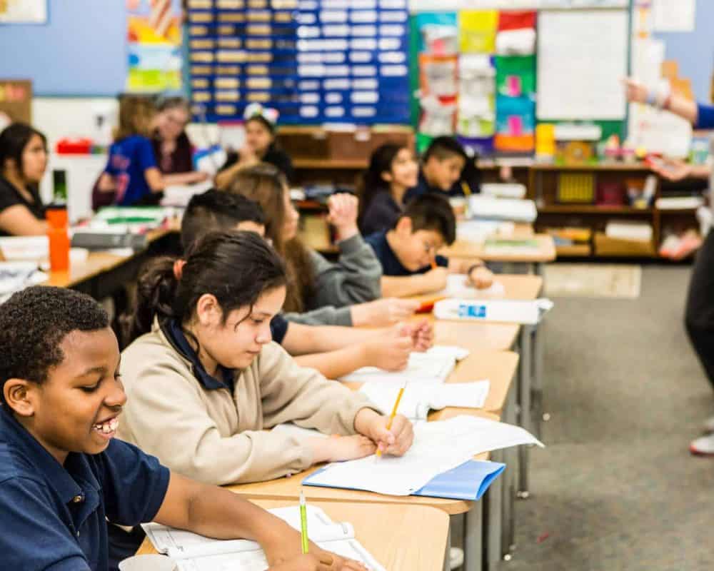 Tulsa students in a row write in notebooks as their teacher gives directions
