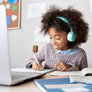 a girl writes in a notebook with headphones on in front a computer