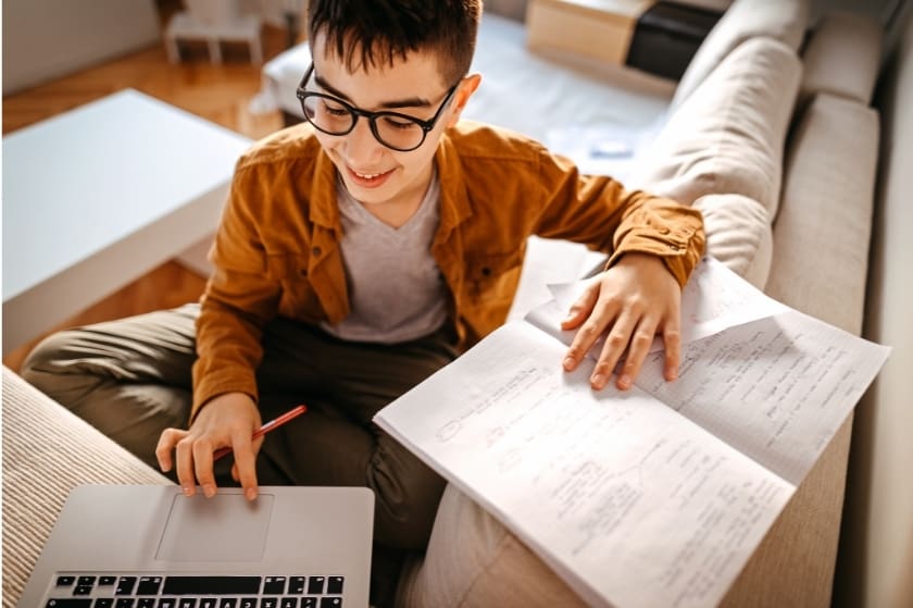 a young boy seated on a couch does work in a notebook while clicking on a laptop