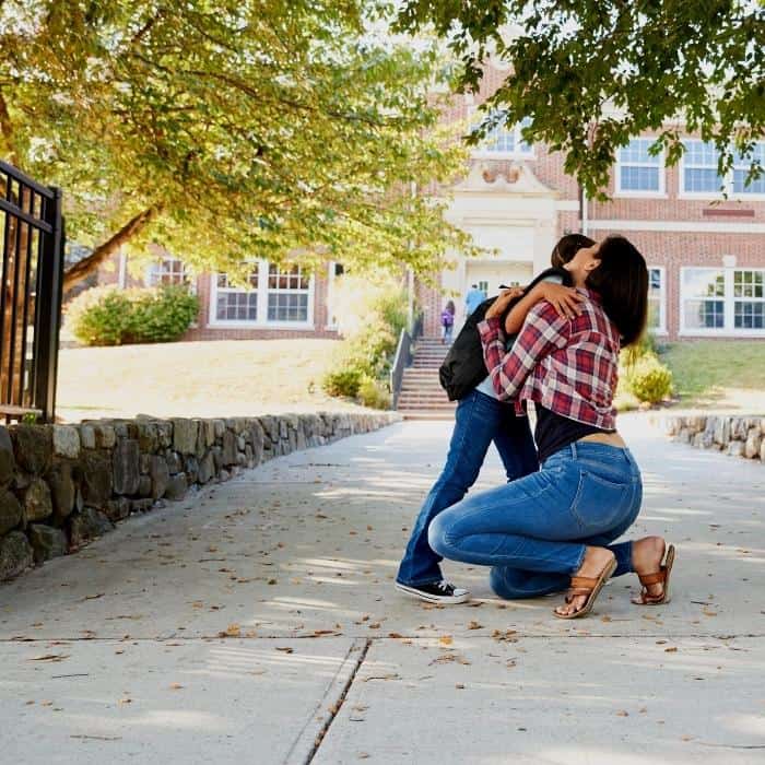 a mother kneels to huge her daughter at drop off outside of a school